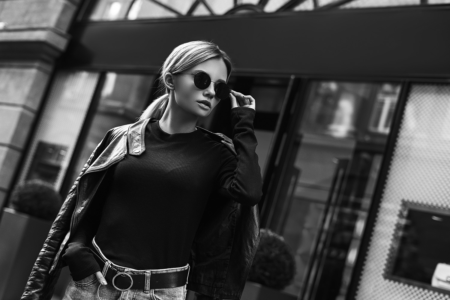 Fashion stylish woman in leather jacket, jeans, sweater and sunglasses walking on road on shops background. Elegant trendy outdoors portrait of pretty girl model on city street