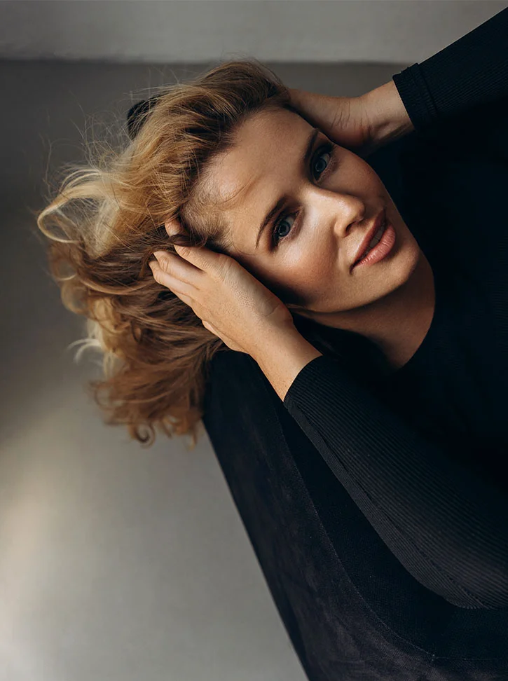 A high-angle close-up shows a light-skinned woman with shoulder-length, wavy light brown hair, hands gently holding her head. She wears a black long-sleeved top and looks slightly upward with a neutral, contemplative expression. Soft lighting and a muted gray background create a calm, intimate mood. - Browlift in New York, NY