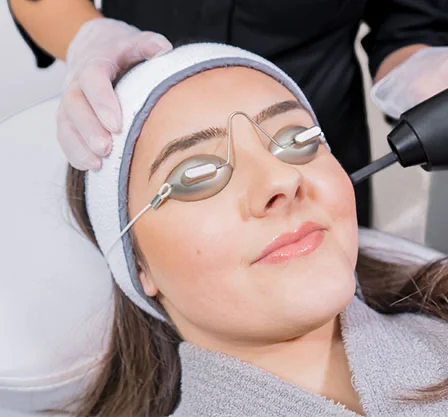 A young woman lies on a spa bed receiving a facial treatment. She wears protective metal eye shields with dark inserts, a light gray headband, and a gray robe. A medical professional in light-colored gloves attends to her, with a dark-colored laser or light therapy device positioned near her face. - Co2 Laser in New York, NY