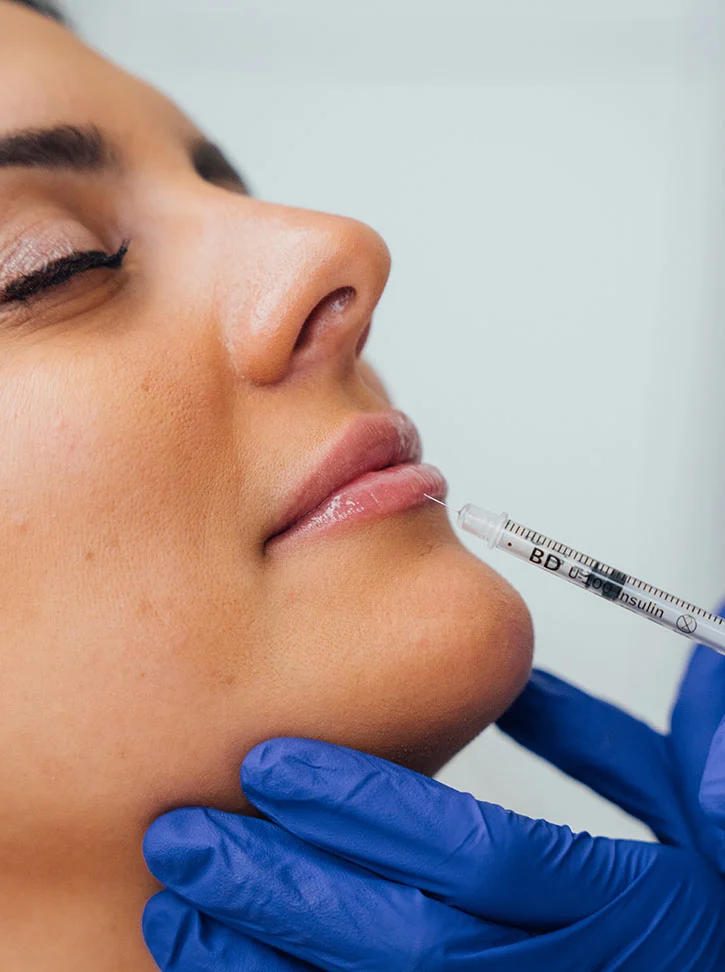 Close-up of a woman receiving a cosmetic injection. Her face is angled slightly upward, eyes closed, with a calm expression. A gloved hand in blue nitrile gloves is seen holding a clear plastic syringe, positioned near her lower lip, ready for injection. The background is light and neutral, keeping the focus on the procedure. - Dermal Fillers in New York, NY