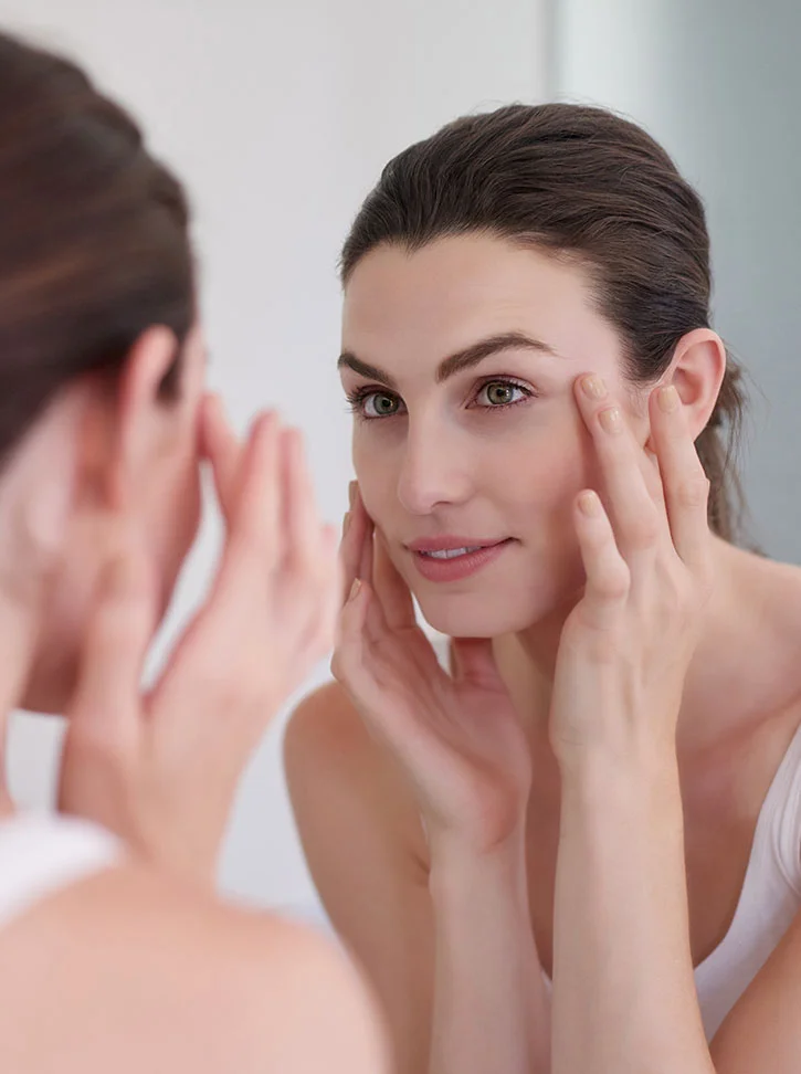 A light-skinned woman with shoulder-length brown hair looks at her reflection in a mirror, gently touching her face with both hands. She wears a light-colored tank top and has a neutral expression, her gaze meeting the camera through the mirror. The softly lit background, likely a bathroom or vanity area, is out of focus, emphasizing the intimate, skincare-focused moment. - Eyelid Surgery in New York, NY