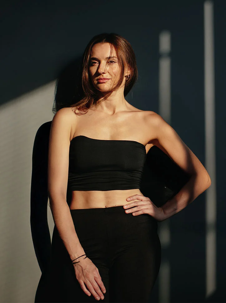 A light-skinned woman with long, light brown hair stands in a studio, bathed in natural light that casts prominent vertical shadows on the wall behind her. She wears a fitted black strapless crop top and matching black pants, with a thin metal bracelet on her left wrist. Her hands rest on her hips, and she gazes neutrally toward the camera. The dark, neutral background and dramatic lighting create depth and highlight her relaxed yet confident posture. - Mommy Makeover in New York, NY
