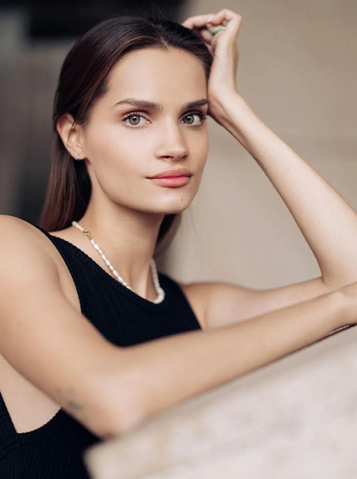 A young woman with shoulder-length brown hair and light tan skin looks directly at the camera with a neutral expression. She has light green or hazel eyes and soft pink lips. She wears a black ribbed tank top and a delicate pearl necklace. Her right arm rests on her head, and her left arm is bent, resting on a surface. The background is a muted beige or light tan. - Morpheus8 in New York, NY
