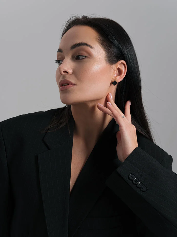 A woman with long dark hair and light skin is shown from the mid-chest up, angled slightly toward the viewer. She wears a fitted black pinstriped blazer and small black clover-shaped earrings. Her expression is relaxed and thoughtful, with her gaze directed off to the side. Light makeup highlights her cheekbones, and her hand gently touches her neck. The background is plain light gray, giving the image a professional, stylish tone. - Necklift in New York, NY