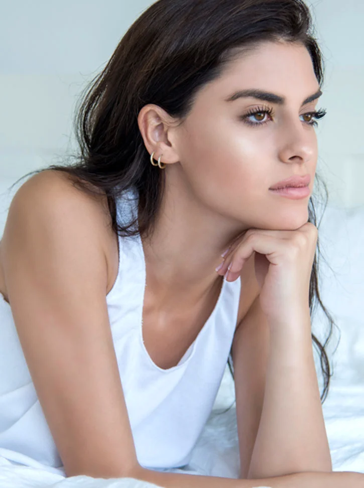 A young woman with long dark hair and light skin lies on a bed, wearing a simple white tank top and small gold hoop earrings. Her head is turned slightly to the right, with a thoughtful expression as she gazes into the distance. One hand rests on her chin, the other along her side. Soft, natural light illuminates her face and body, with a light-gray background and white bedsheets creating a gentle, serene atmosphere. - Otoplasty in New York, NY