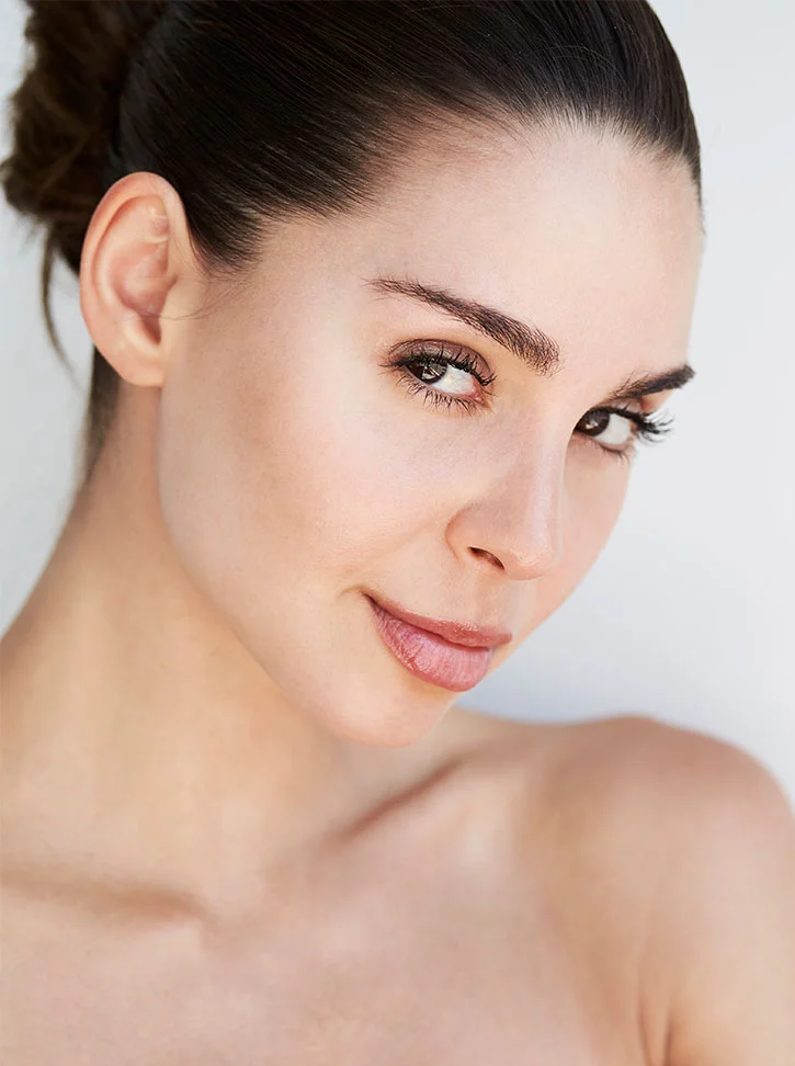Close-up of a woman with shoulder-length dark brown hair pulled back in a bun. She has smooth, light-toned skin, defined eyebrows, and rosy brown lips. Her gaze is directed slightly to the viewer's right. The image highlights her facial features and skin texture with soft lighting against a plain light gray background. - Rhinoplasty in New York, NY